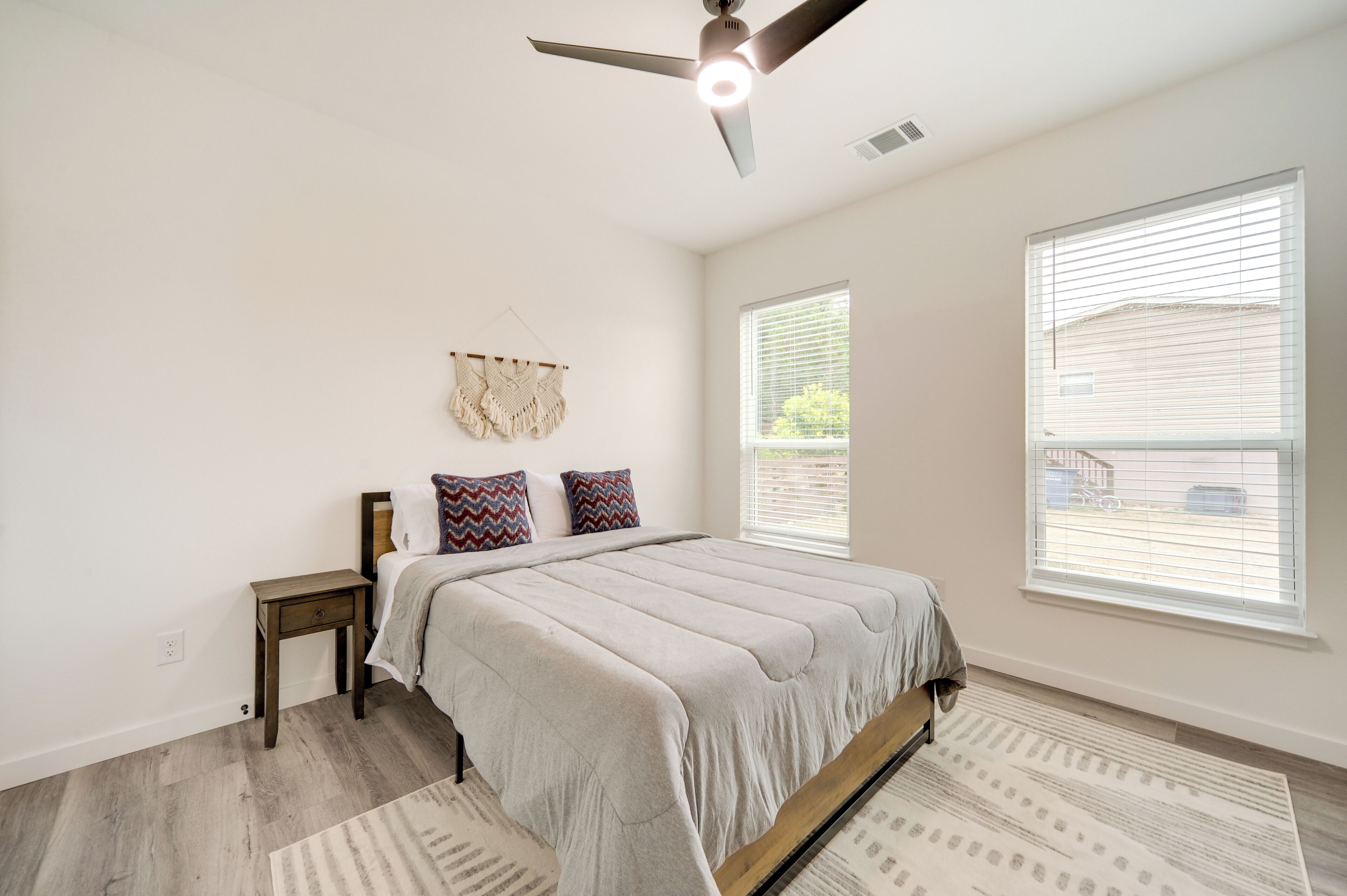 A bedroom with a bed, a ceiling fan, a small bedside table, and two windows with blinds.
