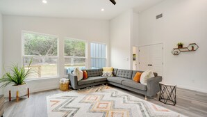 Un salón amplio con un sofá seccional gris, un alfombra estampada y ventanas grandes que ofrecen vistas al exterior.