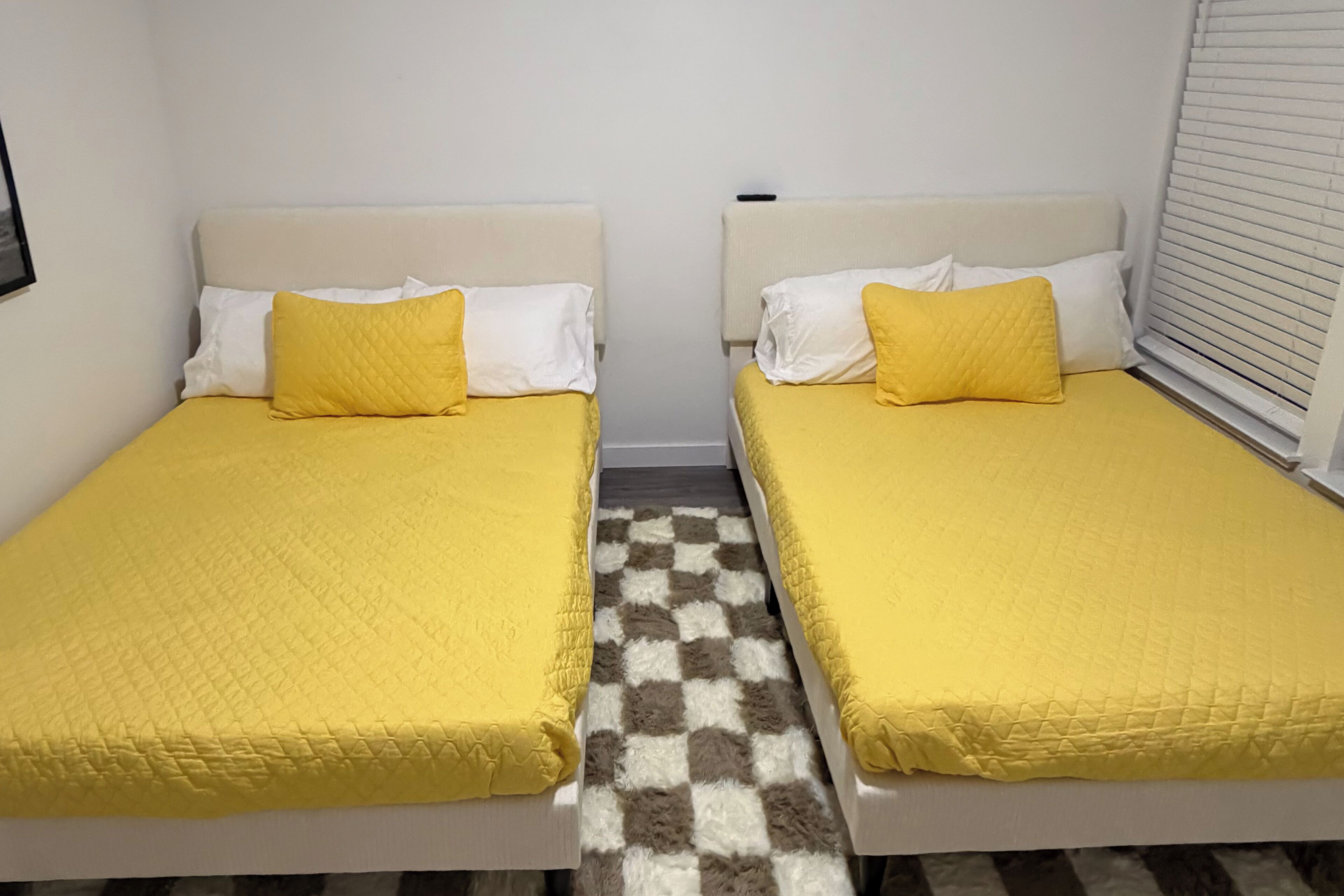 Dos camas individuales con ropa de cama amarilla, un tapete a cuadros y una ventana con persianas.