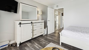 A bedroom with a white bed, a dresser with drawers, a TV mounted on the wall, and a door leading to another room. - Beachfront Fourth-floor Condo w/ Community Pool! (North Wildwood)