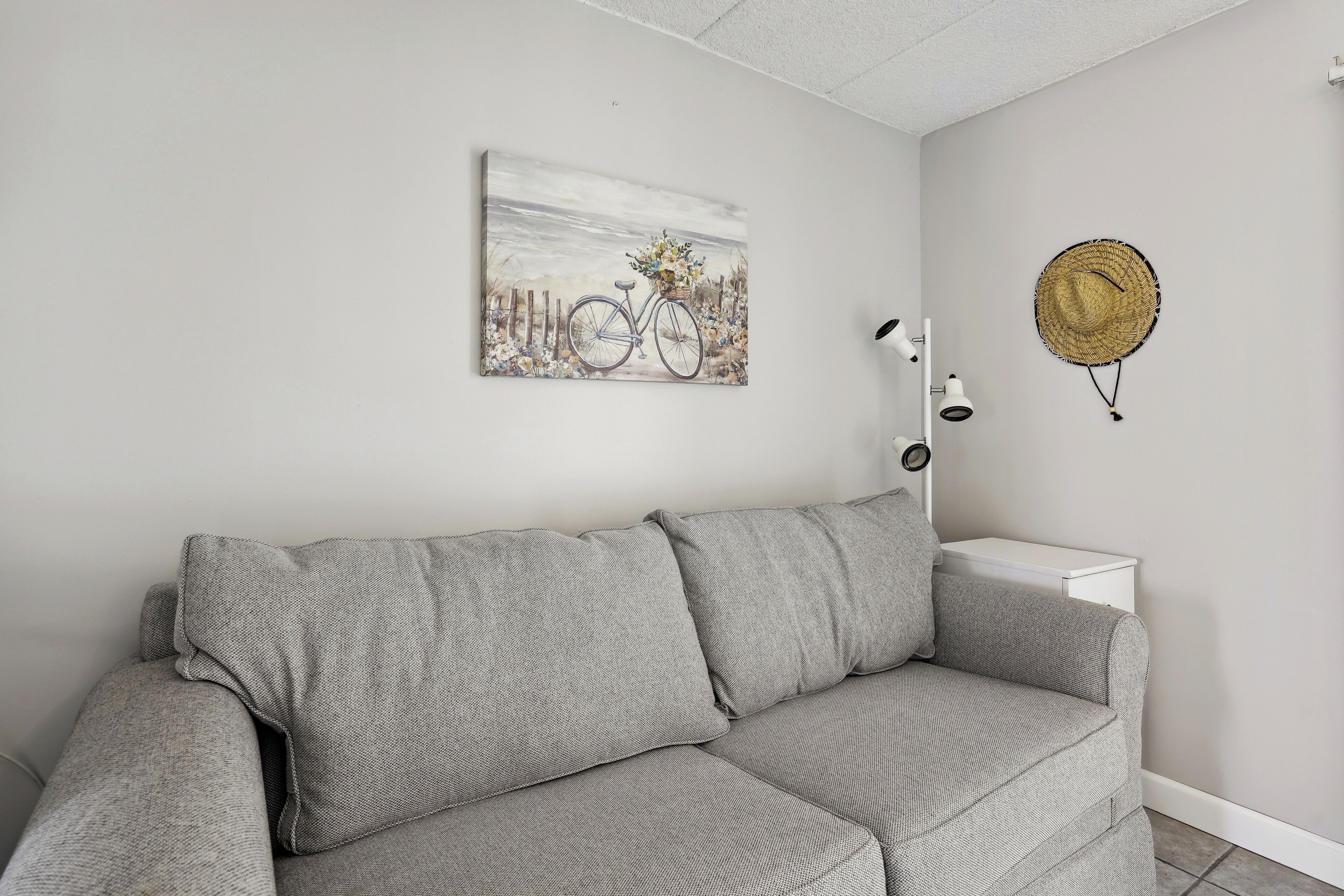 Un canapé d’angle gris est placé contre un mur blanc, et on y voit un tableau encadré représentant un vélo ainsi qu’un panier en osier.