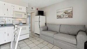 A compact kitchen with white cabinets, a microwave, a stove, and a refrigerator. A gray sofa is positioned against the wall, and a painting of a bicycle is displayed above it. - Beachfront Fourth-floor Condo w/ Community Pool! (North Wildwood)