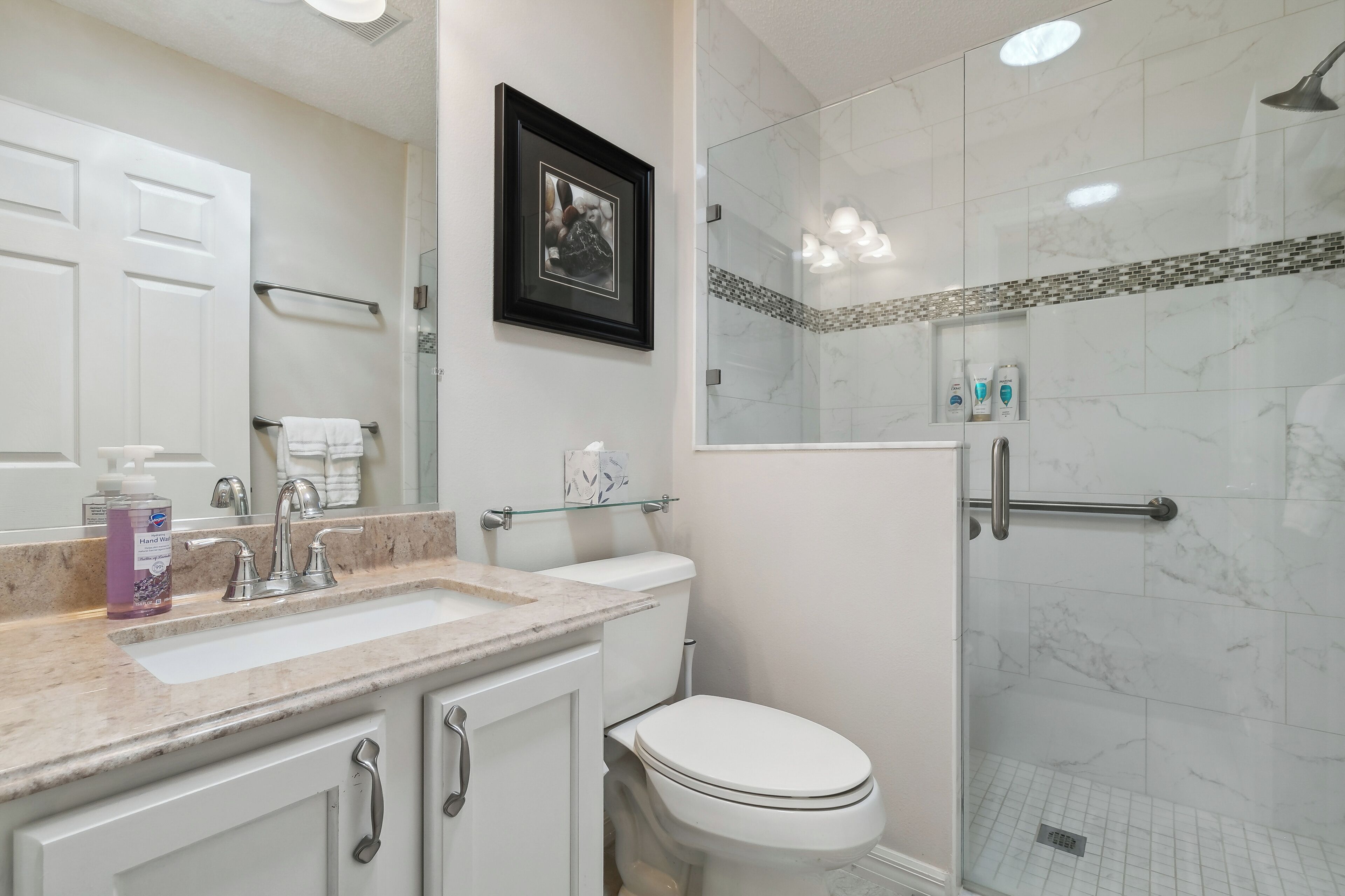 A modern bathroom with a marble sink, a framed picture, a glass shower enclosure, and a white toilet.