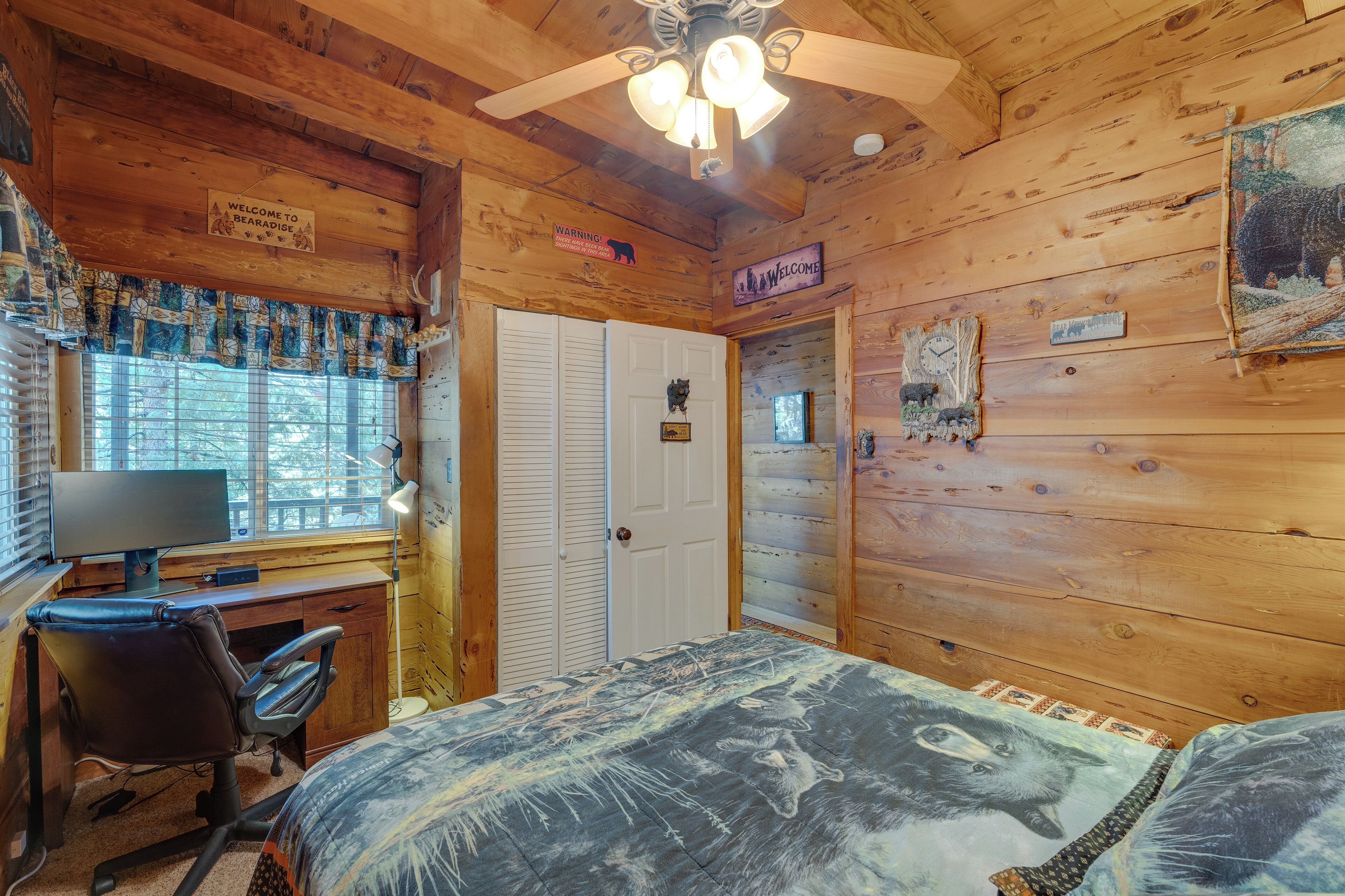 L’intérieur d’une cabane avec un plafond en bois, un lit recouvert d’une couverture à motifs d’ours, un bureau avec un ordinateur et une fenêtre avec des stores.