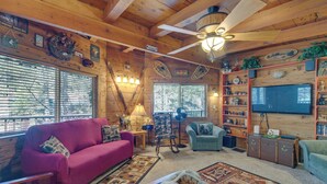 L’intérieur chaleureux d’un chalet avec un plafond en bois, un ventilateur de plafond, un téléviseur, une cheminée et une grande fenêtre équipée de stores.