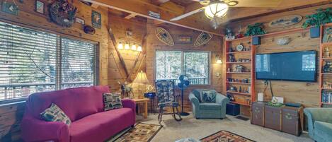 A cozy cabin interior with a wooden ceiling, a ceiling fan, a television, a fireplace, and a large window with blinds.