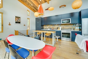 A modern kitchen with dark cabinets, a stainless steel oven, and a microwave. There is a dining table with blue and red chairs.