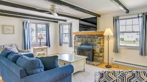 A cozy living room with a blue sofa, a glass coffee table, a stone fireplace, and a television mounted above it. - Cozy Westbrook Vacation Rental - Walk to Beach! (Westbrook)