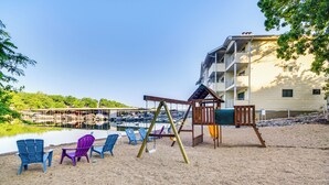 A sandy beach area with a swing set and colorful Adirondack chairs. In the background, there is a building with multiple balconies and a dock with boats.
