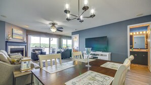 A dining area with a wooden table, white chairs, and a chandelier. There is a fireplace with a mantel and a television on the wall.