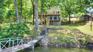 A two-story wooden cabin with a deck and stairs leading to a dock over a body of water.