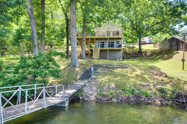A two-story wooden cabin with a deck and stairs leading to a dock over a body of water.