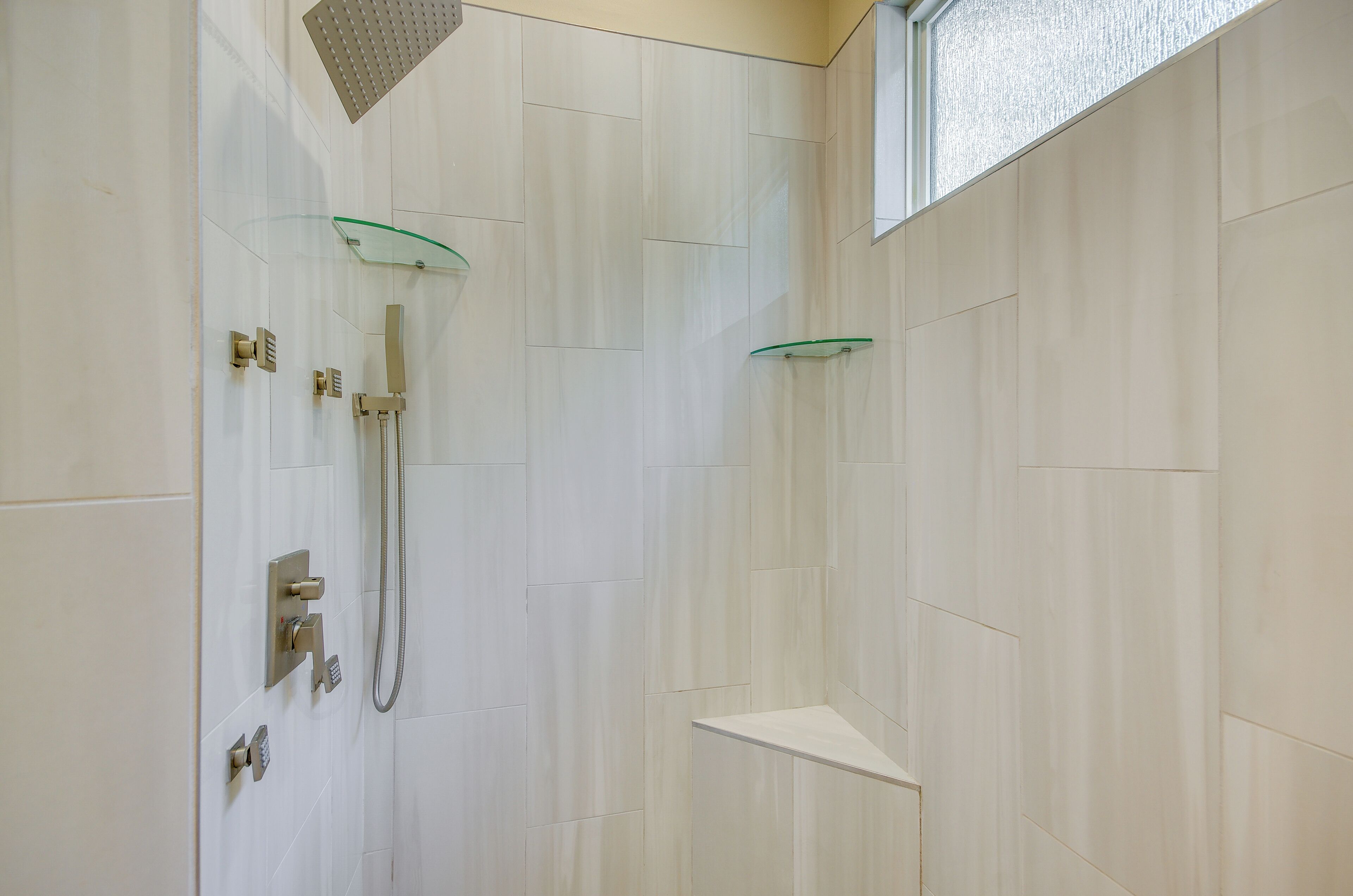 A shower with a glass shelf, a handheld showerhead, and a wall-mounted control panel.
