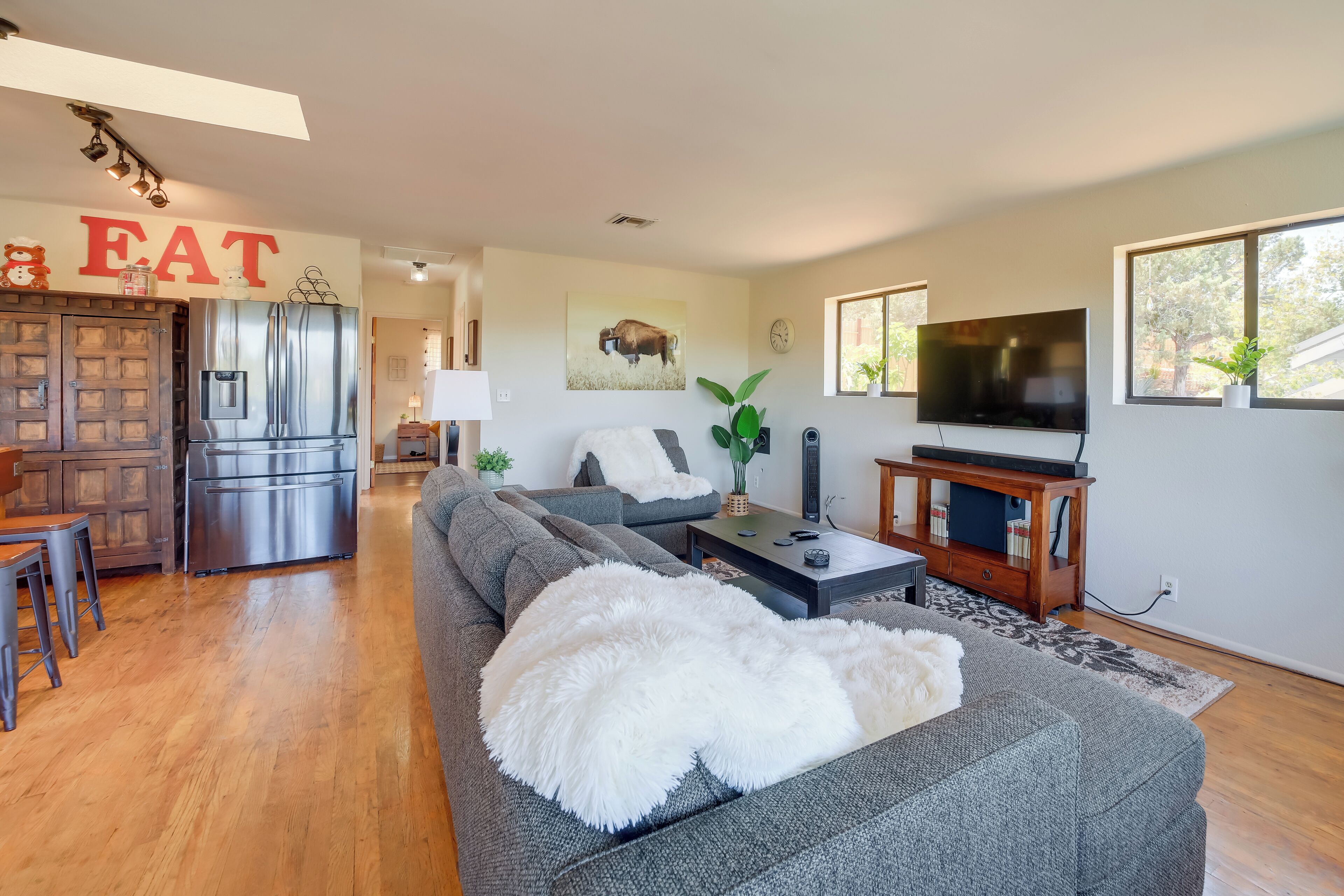 A modern living room with a grey sofa, a white fluffy throw, a TV, and a kitchen area with a stainless steel refrigerator.