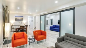 Ein modernes Wohnzimmer mit einem grauen Sofa, zwei orangefarbenen Sesseln und einem blauen Schlafsofa. Es gibt eine Essecke mit Tisch und Stühlen sowie ein großes Fenster mit Vorhängen.