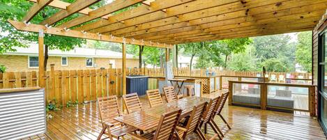 A covered outdoor deck with a wooden table and chairs, surrounded by a wooden fence and a view of a house in the background.