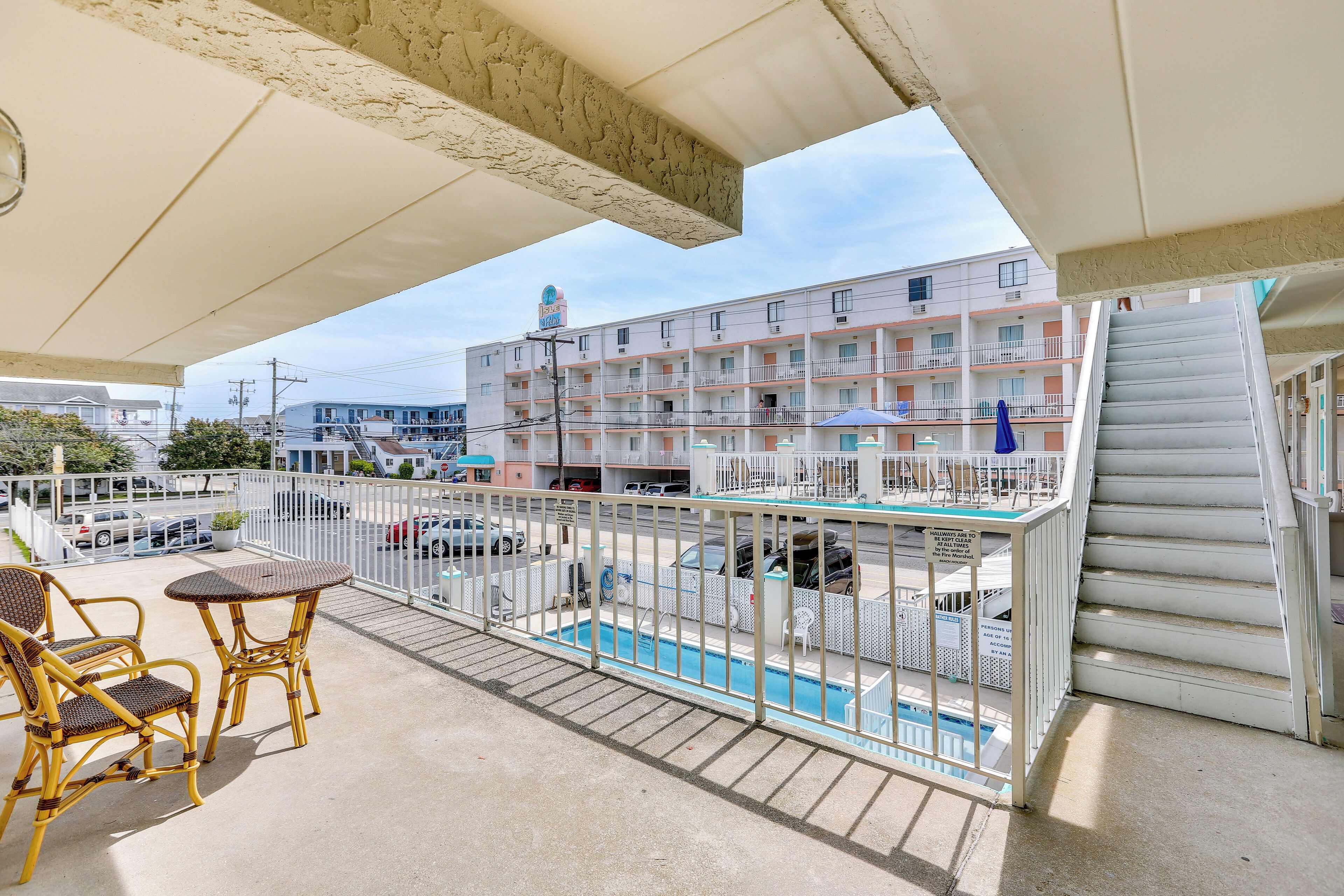 A balcony with wicker chairs and a table, overlooking a swimming pool and a multi-story building with balconies.