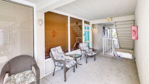 A covered outdoor seating area with wicker chairs and a small table, a staircase leading to an upper level, and a visible exit sign. - Wildwood Condo w/ Community Pool: Walk to Beach! (Wildwood)