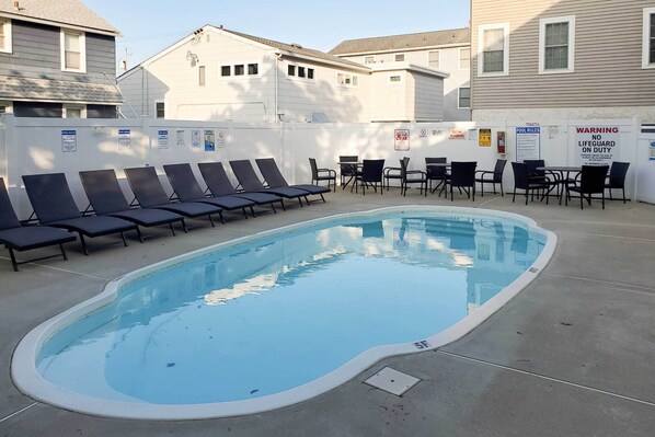Piscina com espreguiçadeiras e mesas, cercada por um muro branco com várias placas.