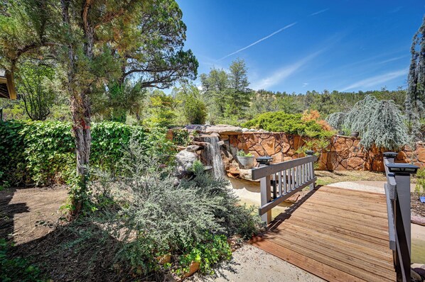 Una terraza de madera que conduce a un jardín de rocas con cascada, rodeado de diversos árboles y arbustos.