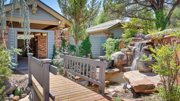 Une terrasse en bois menant à une maison à l’extérieur en briques, avec une petite cascade et une végétation luxuriante tout autour.