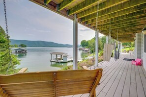 Une chaise suspendue en bois, installée sur une terrasse couverte qui donne sur un lac où l’on peut voir des bateaux et des quais.