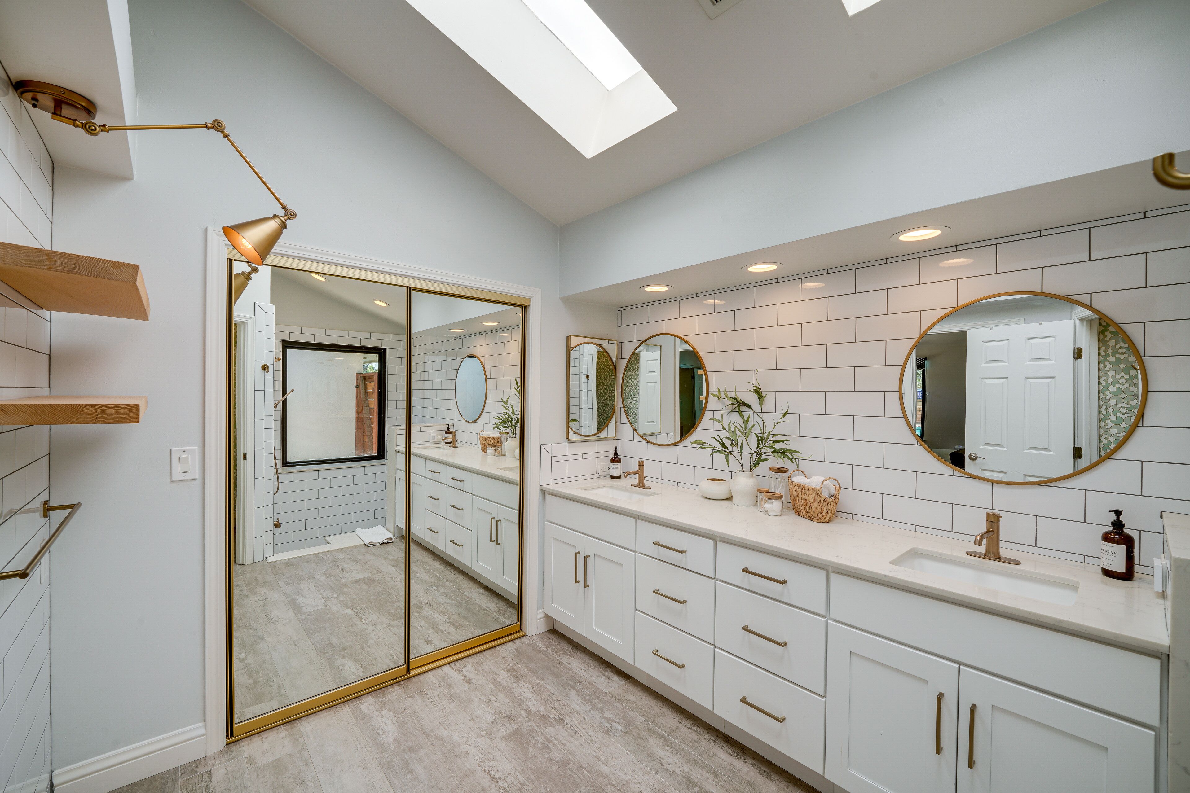 A modern bathroom with white tiled walls, a skylight, a large mirror, and a vanity with a sink and multiple drawers.