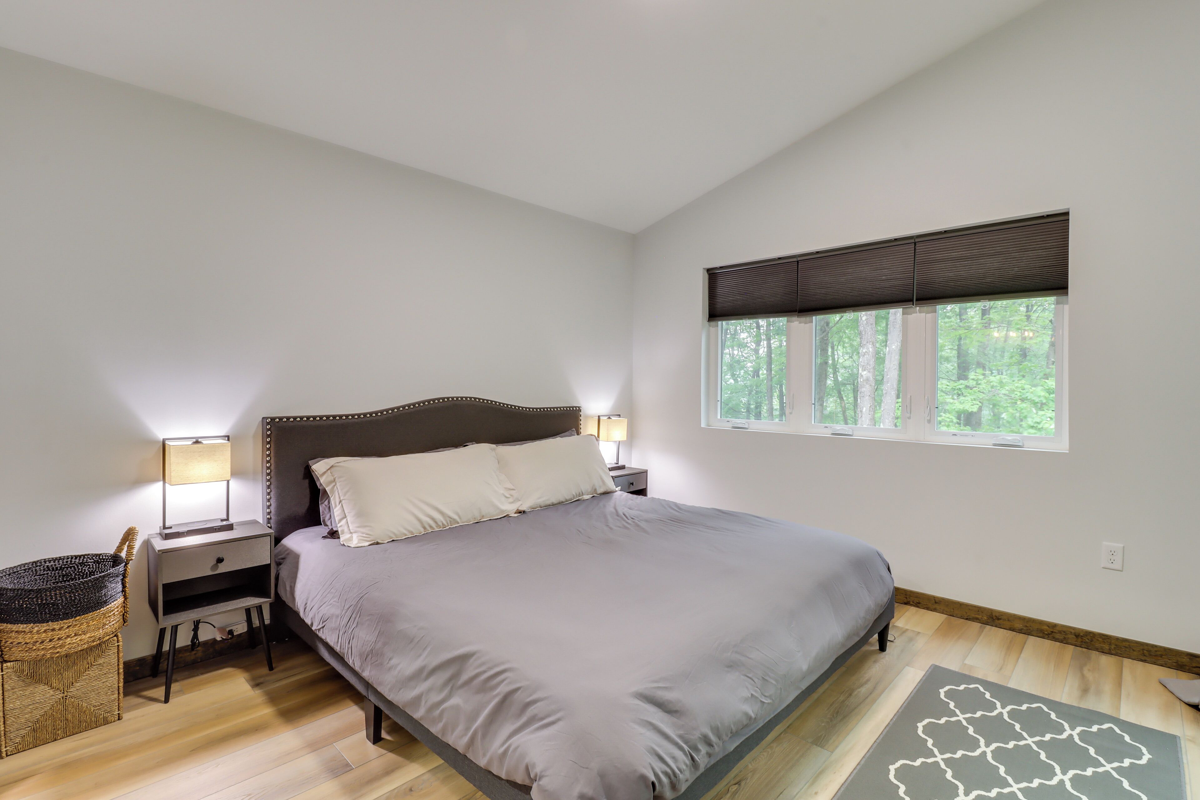 Quarto com cama, mesas de cabeceira, abajur, janela e vista para árvores.