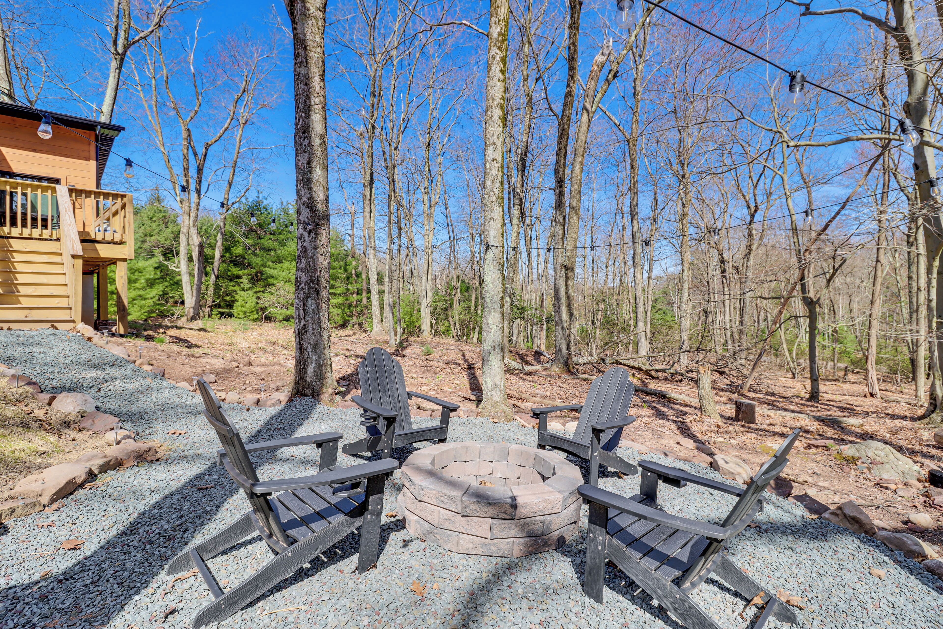 A wooden deck with Adirondack chairs and a stone fire pit surrounded by gravel, set against a backdrop of trees and a wooden cabin.