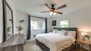 Un dormitorio con cama, ventilador de techo, mesita de noche, cómoda y dos ventanas.