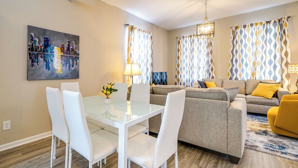 Una sala de estar moderna con una mesa de comedor de vidrio, sillas blancas, un sofá gris y cojines amarillos.
