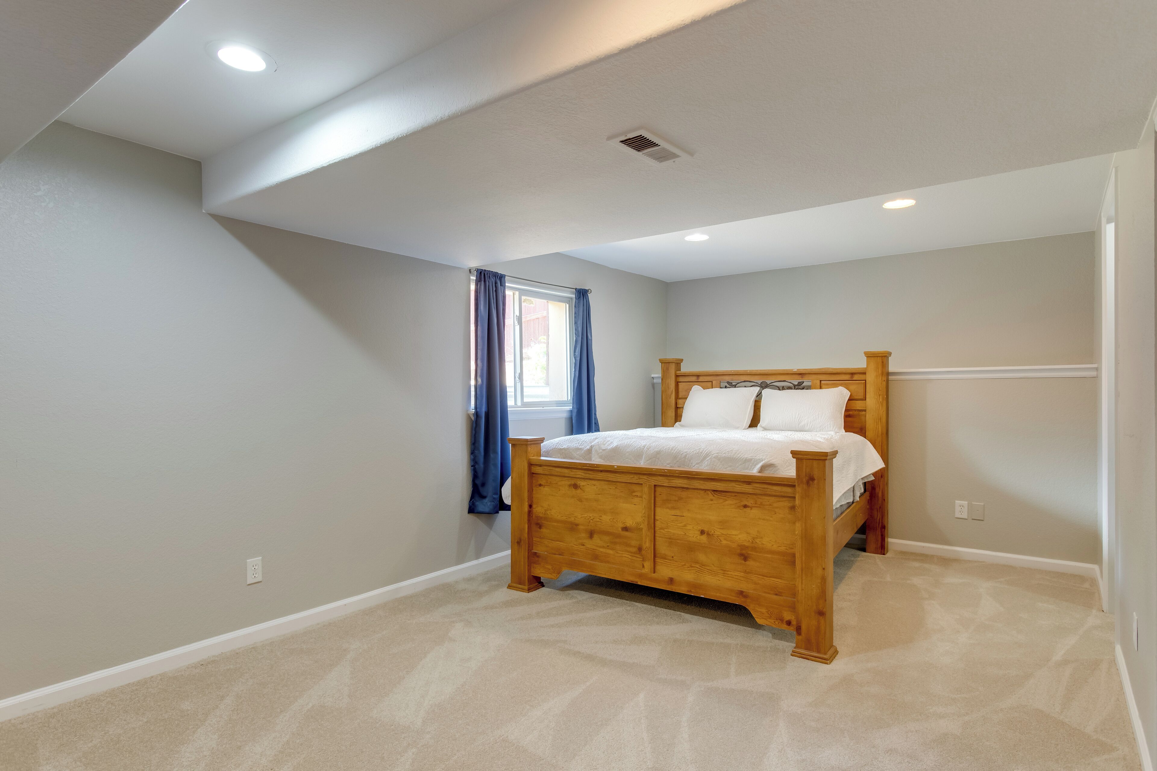 A bedroom with a wooden bed, a window with blue curtains, and a sloped ceiling with recessed lighting.