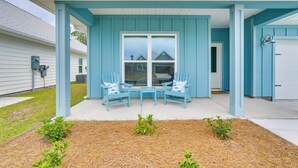 Varanda coberta com duas cadeiras azuis e uma mesa pequena, porta de garagem branca e uma casa com revestimento azul.