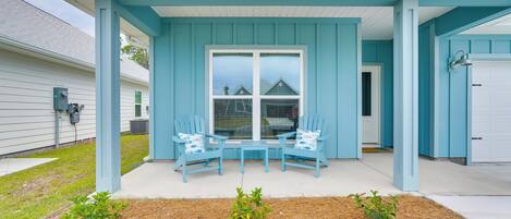 Un porche couvert avec deux chaises bleues et une petite table, une porte de garage blanche et une maison aux bardages bleus.
