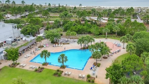 Piscina cercada por palmeiras e cadeiras de praia, com vista para a praia e oceano ao fundo.