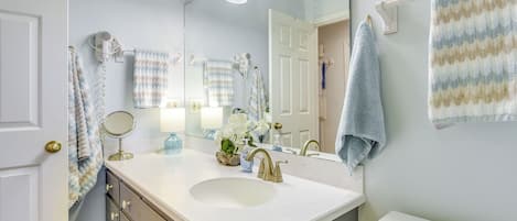 A bathroom with a white sink, gold faucet, and a large mirror. There are multiple towels hanging on the wall and a hairdryer on the wall.
