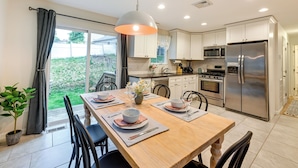 A modern kitchen with white cabinets, stainless steel appliances, and a dining area with a wooden table set for two. - Walk to Shops & Dining: Glen Cove Apt w/ Yard! (Glen Cove)