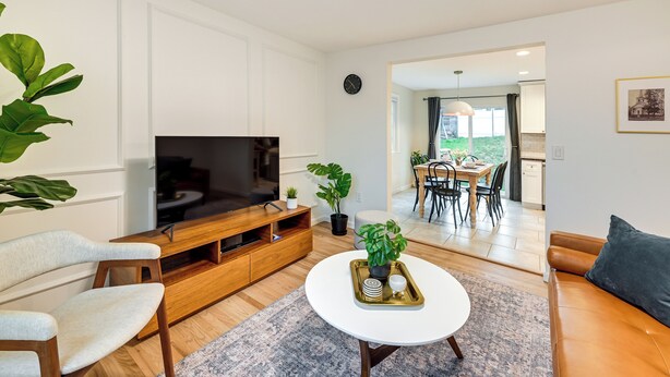 A modern living room with a flat-screen TV, wooden furniture, a white coffee table, and a dining area visible through an open doorway. - Walk to Shops & Dining: Glen Cove Apt w/ Yard! (Glen Cove)