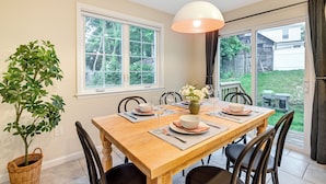 A dining area with a wooden table, chairs, a hanging light fixture, and a view of the backyard through large windows. - Walk to Shops & Dining: Glen Cove Apt w/ Yard! (Glen Cove)