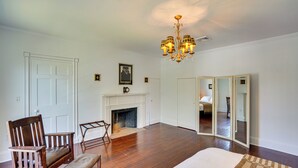 A spacious room with hardwood flooring, a large bed, a wooden chair, a bench, a fireplace, and a chandelier. - Coxsackie Home w/ River Views: Walk to The Wire! (Coxsackie)