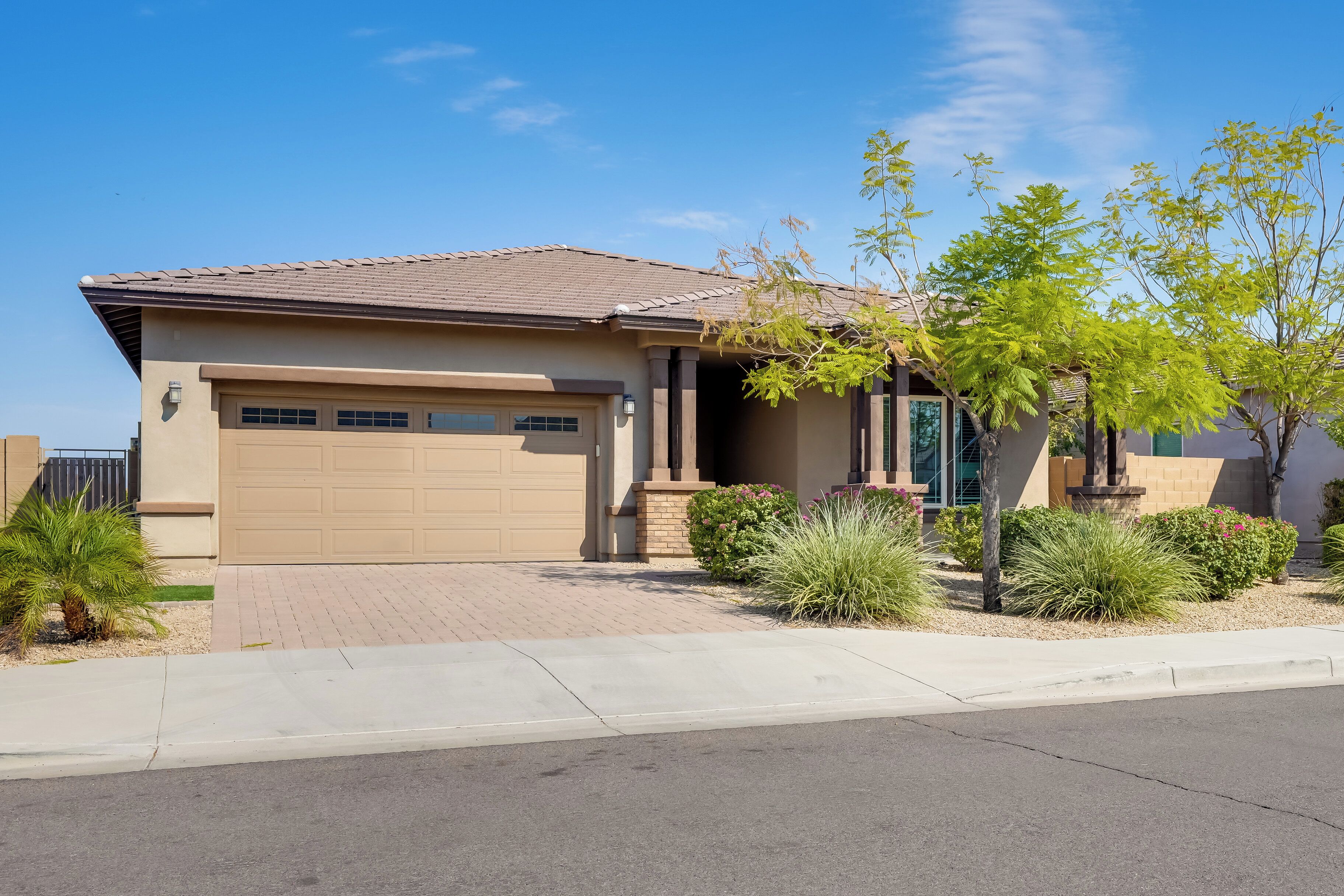 A single-story house with a beige garage door, a tiled roof, and a landscaped front yard with various shrubs and a small tree.