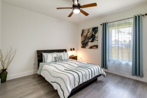 Une chambre à coucher avec un lit, un ventilateur de plafond, une table de chevet, une lampe, une fenêtre avec des rideaux et un tableau au mur.