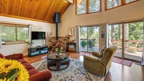 A spacious living room with a high ceiling, a fireplace, a TV, and a large window overlooking a deck. - Groveland Vacation Rental ~ 26 Miles to Yosemite! (Groveland)