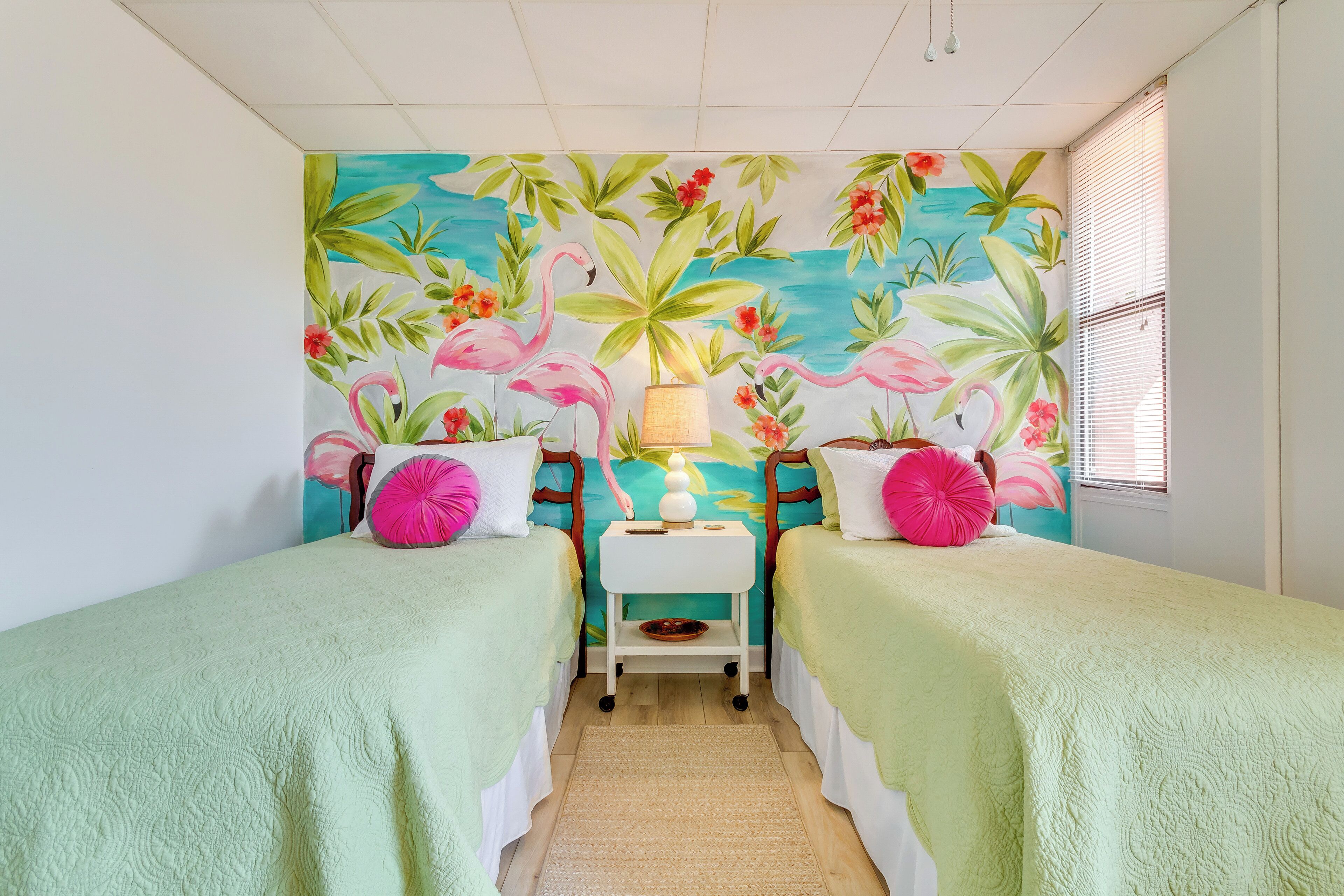 Una habitación con dos camas, una mesita con una lámpara, y un mural con plantas tropicales y flamencos.