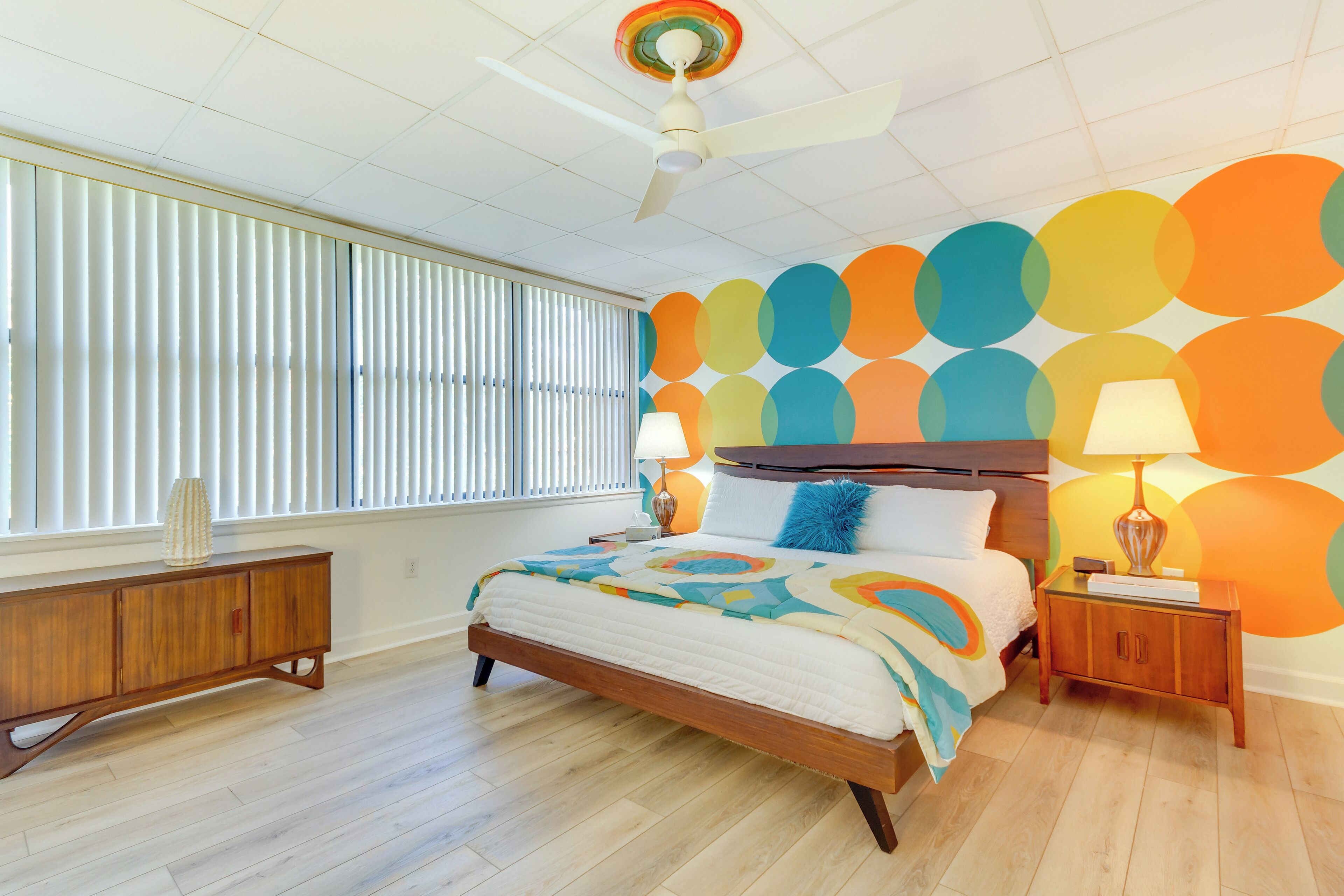 Een slaapkamer met een houten bed, nachtkastje, plafondventilator en een muur met kleurrijke cirkelvormige patronen.