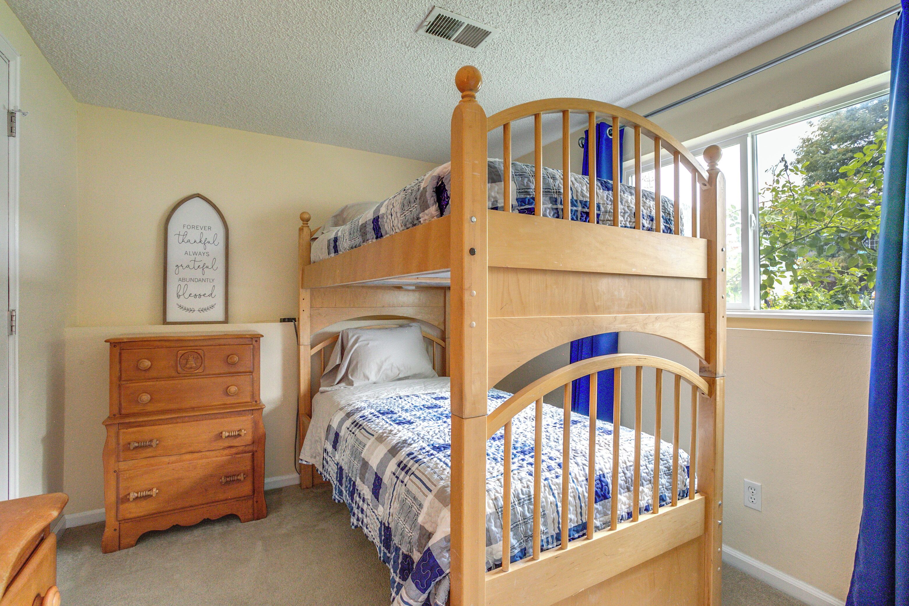 A bunk bed with a wooden headboard, a dresser with drawers, a framed inspirational quote on the wall, and a window with a view of greenery.