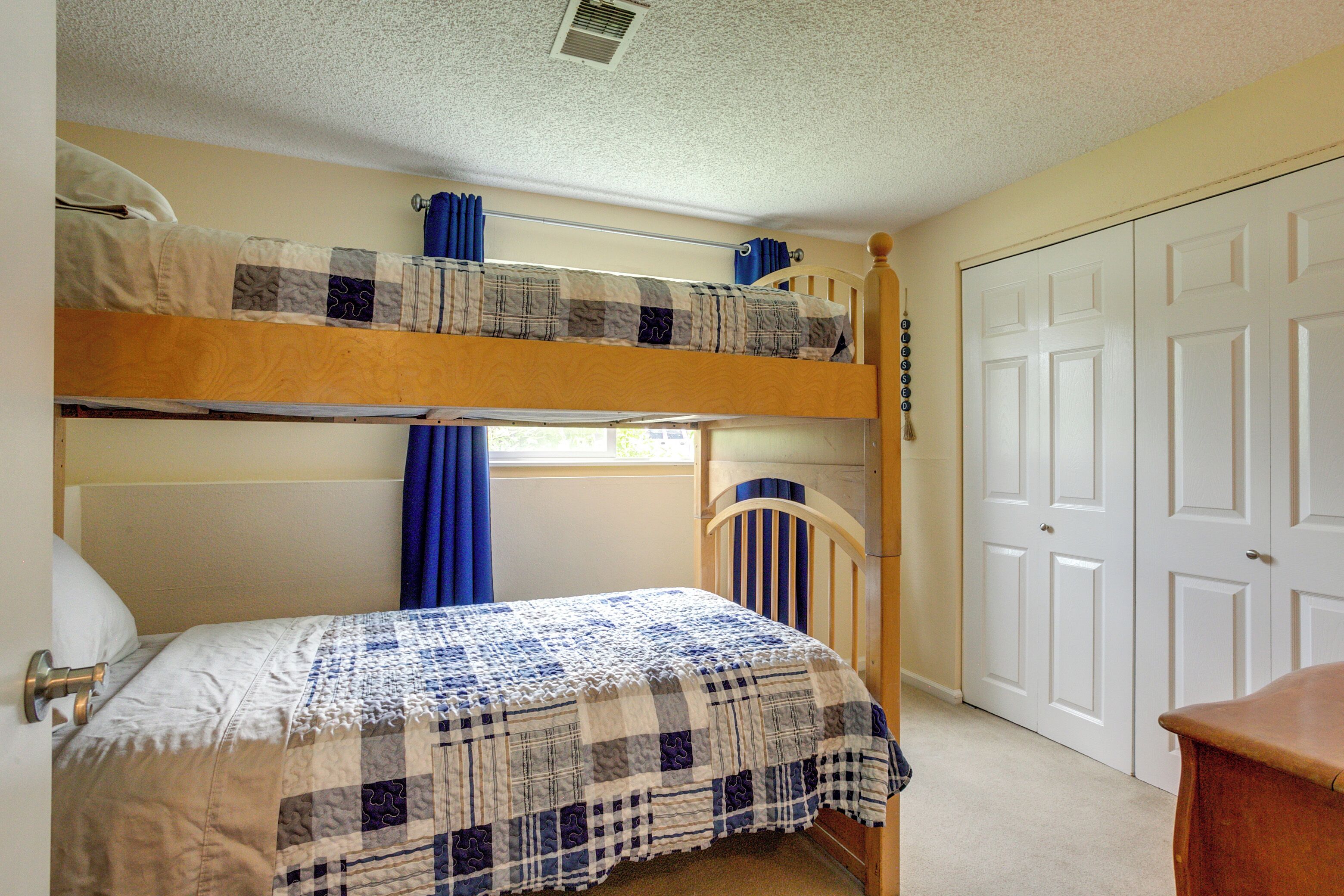 A bunk bed room with a checkered bedspread, a window with blue curtains, and a white door.