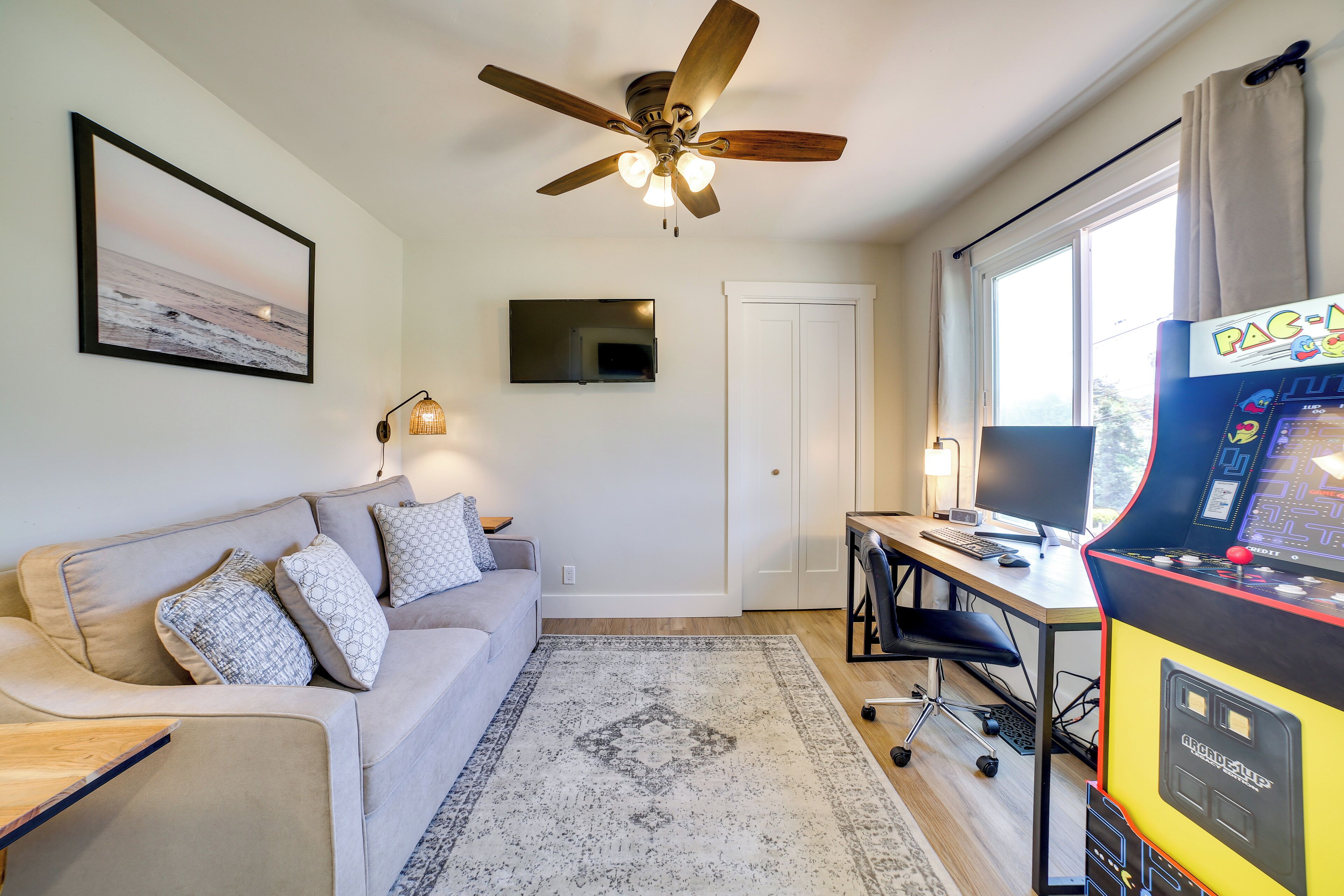 A living room with a ceiling fan, a couch, a TV, a gaming console, and a large window.