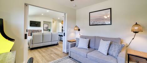 A modern living room with a grey sofa, patterned rug, ceiling fan, and a kitchen area visible through an open doorway.