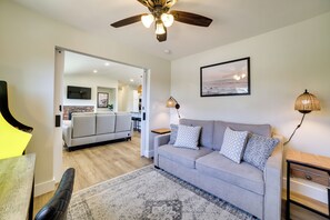 Un salon moderne avec un canapé gris, un tapis à motifs, un ventilateur de plafond et un coin cuisine visible à travers une porte ouverte.